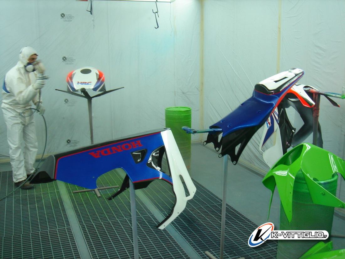Cabina di verniciatura con carene Honda blu e bianche e un parafango verde appesi ai supporti, un verniciatore in tuta sullo sfondo